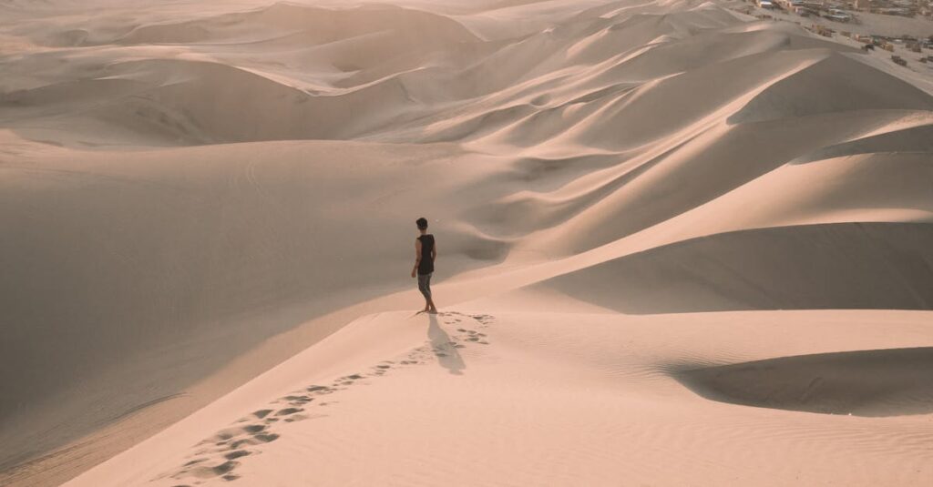A lone traveler walks across the vast sand dunes of Ica, Peru, under the warm glow of a sunset.