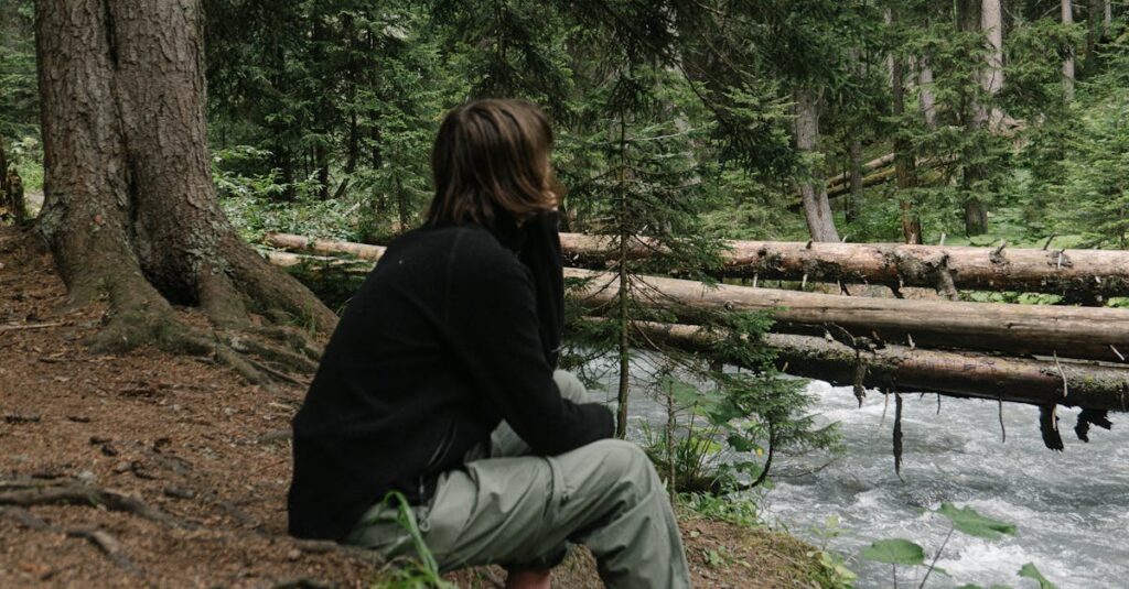 A person in a forest sits by a flowing river, immersed in nature's tranquility.