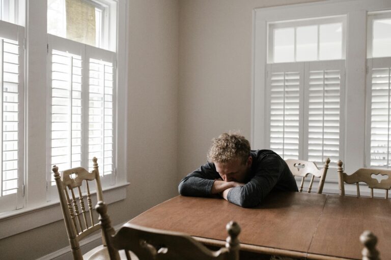 An adult man with emotions of sadness and anxiety sitting alone at a wooden table near windows.