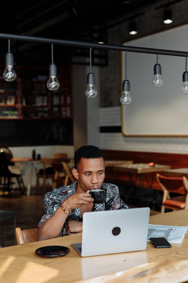 Young man at coffee shop working remotely on laptop, sipping coffee.